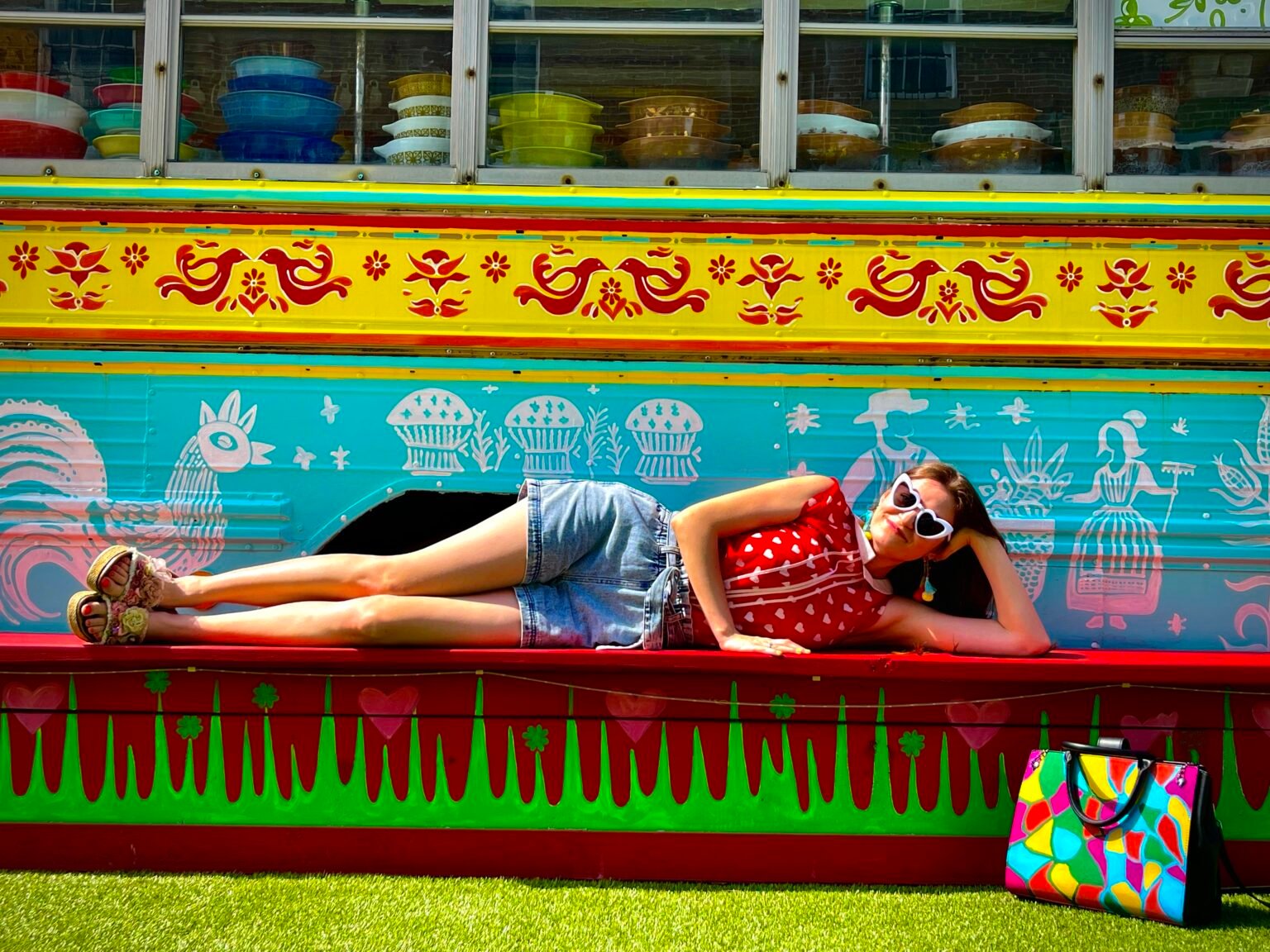 girl wearing colorful outfit, red shirt and high-waisted shorts, laying on a red bench, colorful bus behind her, multicolored bag next to her