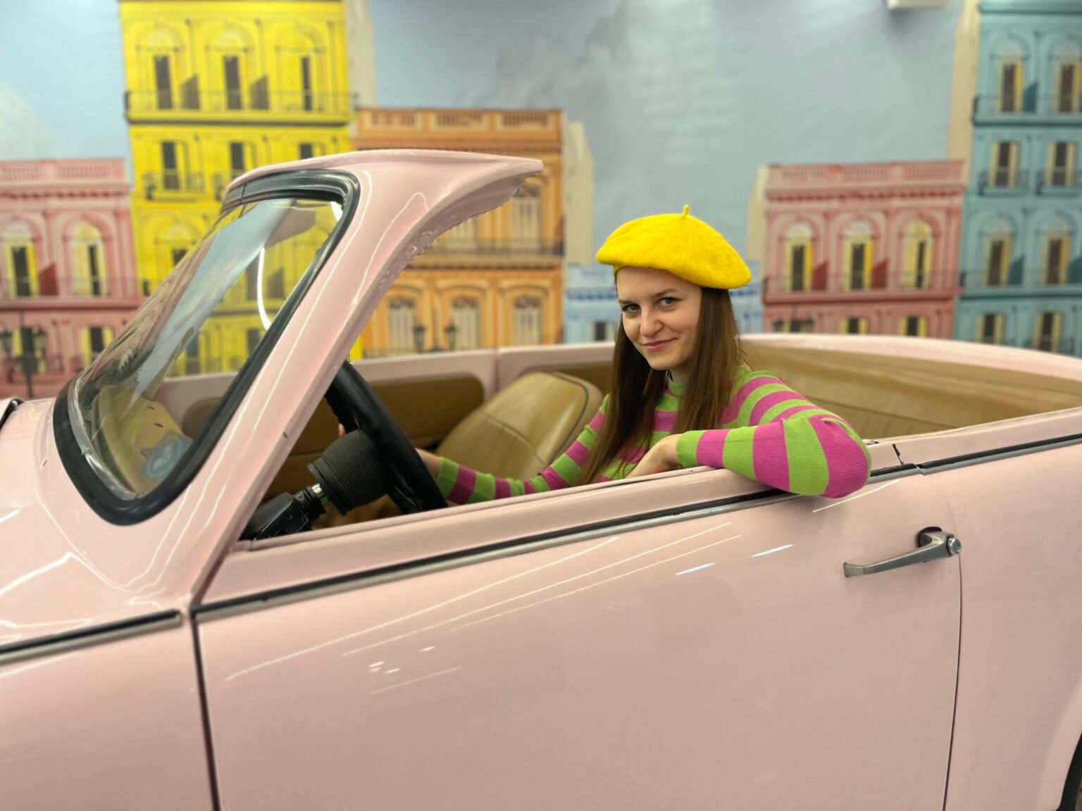 a girl in colorful outfit - yellow beret and pink and green shirt - is driving a pastel pink car, colorful buildings behind her