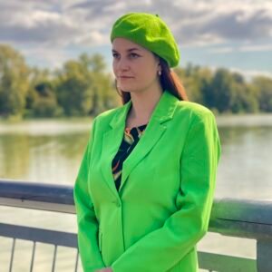 bright green beret side view on a model, she is wearing vibrant green blazer and looking at the horizon, a river and green trees in the background