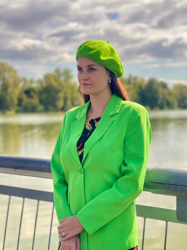 bright green beret side view on a model, she is wearing vibrant green blazer and looking at the horizon, a river and green trees in the background