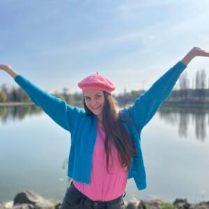 woman wearing candy pink beret, pink shirt and blue cardigan smiling and putting her hands up in the air, a lake in the background