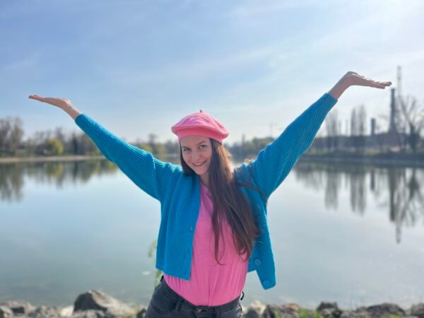 woman wearing candy pink beret, pink shirt and blue cardigan smiling and putting her hands up in the air, a lake in the background