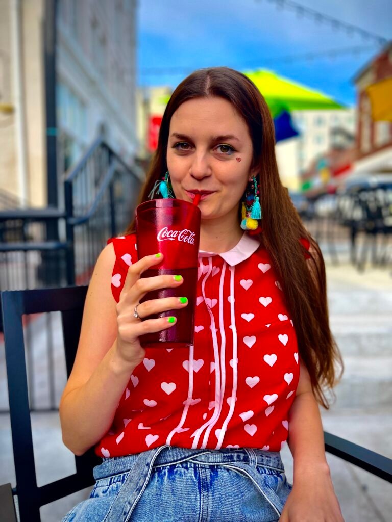 a girl wearing cherry red top with white hearts, a pair of jeans shorts and colorful earrings, she is drinking cola