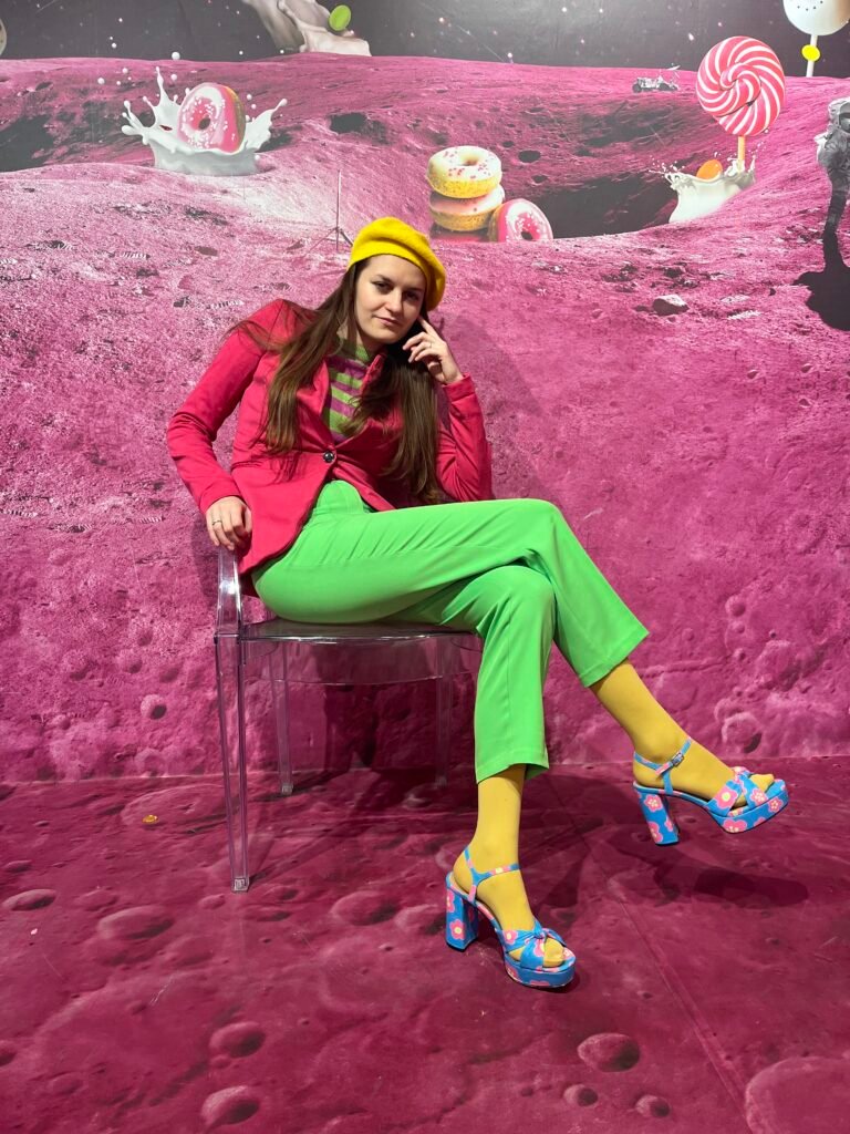 a girl sitting on a transparent chair, abstract pink wallpaper behind her, she is wearing multicolored suit - pink and green, a yellow beret, yellow tights and blue heels.