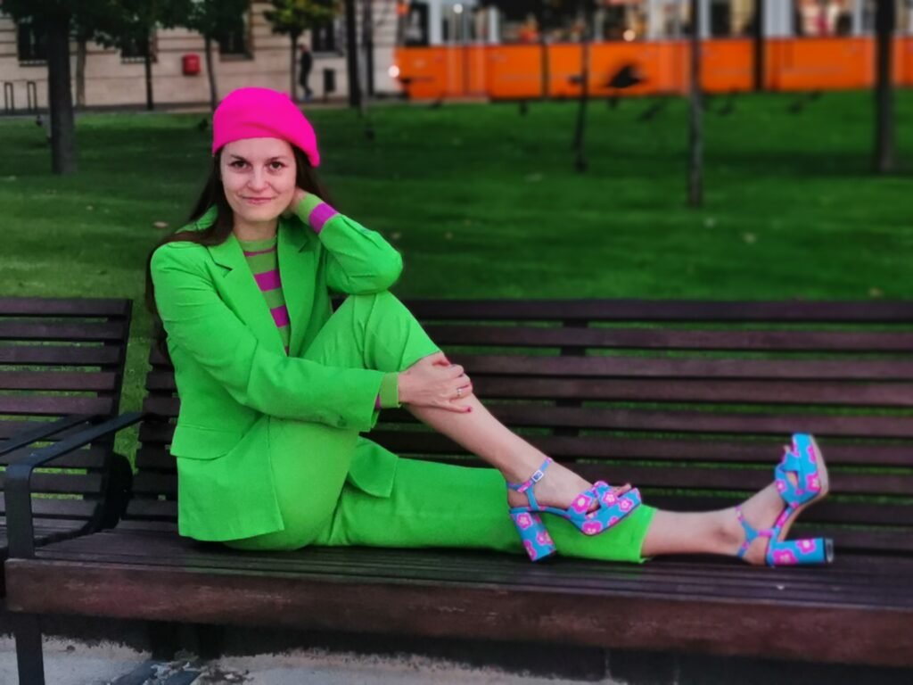 a model wearing colorful suits in a vivid green color, fuchsia beret and blue high heels, sitting on a bench