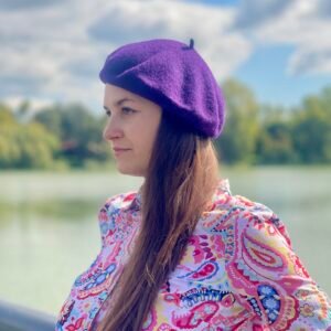 dark purple beret side view on a model wearing colorful button shirt, a river and clouds in the background