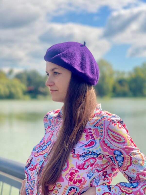 dark purple beret side view on a model wearing colorful button shirt, a river and clouds in the background