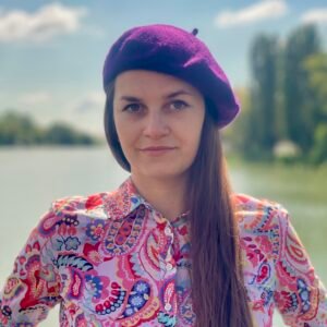 dark purple beret front view on a model, she is wearing colorful button shirt and she has brown hair, a river and trees in the background