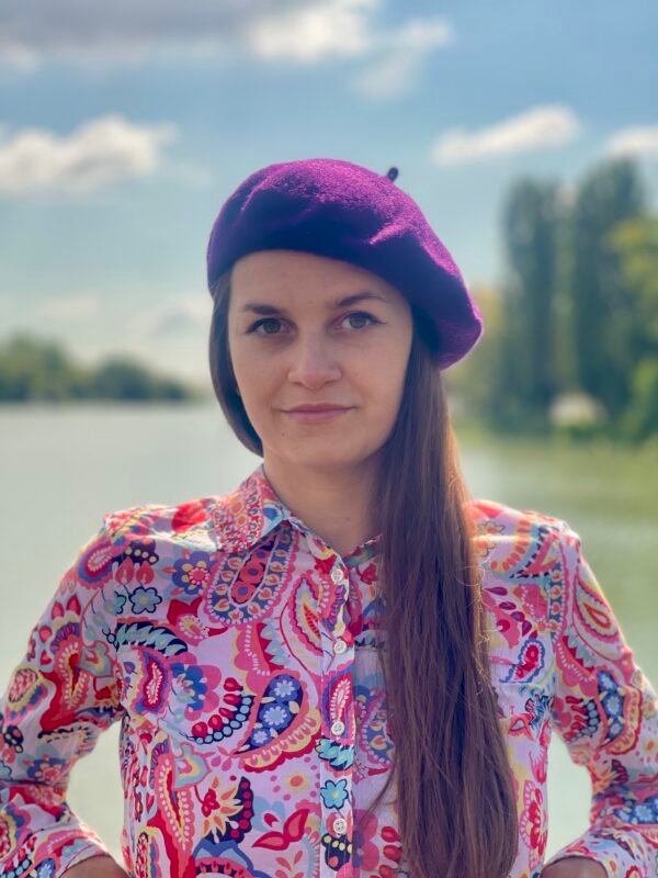 dark purple beret front view on a model, she is wearing colorful button shirt and she has brown hair, a river and trees in the background