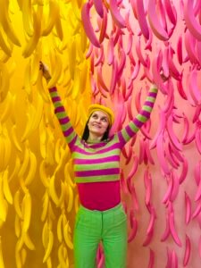 a girl wearing colorful clothes, pink and green striped top, green pants, pink and yellow bananas hanging in the background