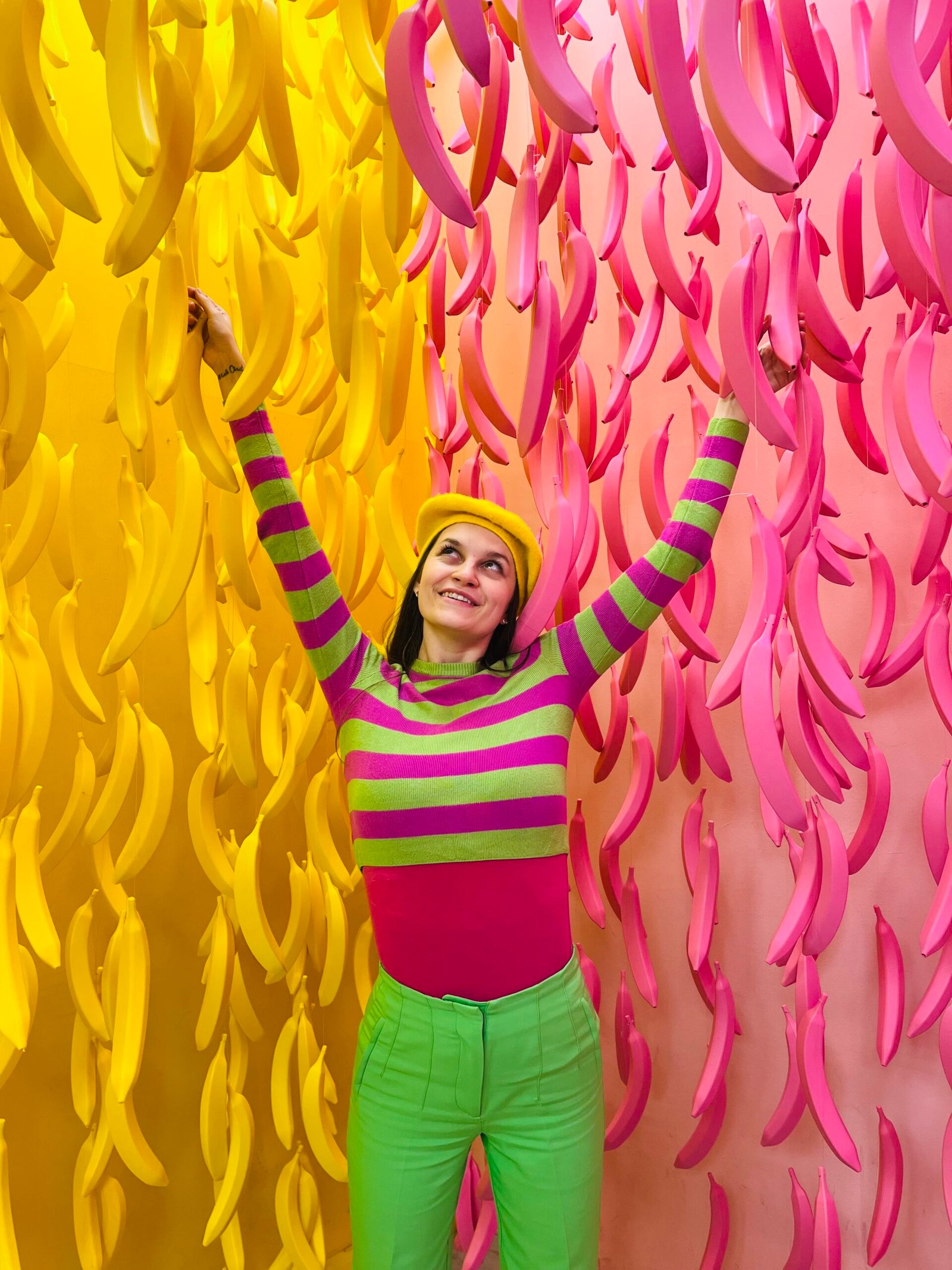a girl wearing colorful clothes, pink and green striped top, green pants, pink and yellow bananas hanging in the background