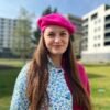 woman wearing fuchsia beret and pink and blue coat, grass and building in the background