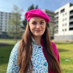 fuchsia beret side view, on a model wearing pink and blue coat, grass and building in the background