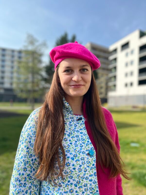 woman wearing fuchsia beret and pink and blue coat, grass and building in the background