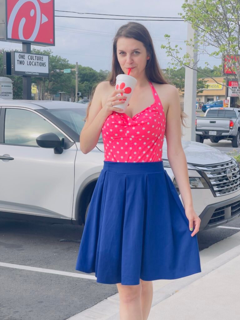 a girl wearing a colorful skirt in navy blue shade, and a pink polka dot top, drinking a drink, a car in the background