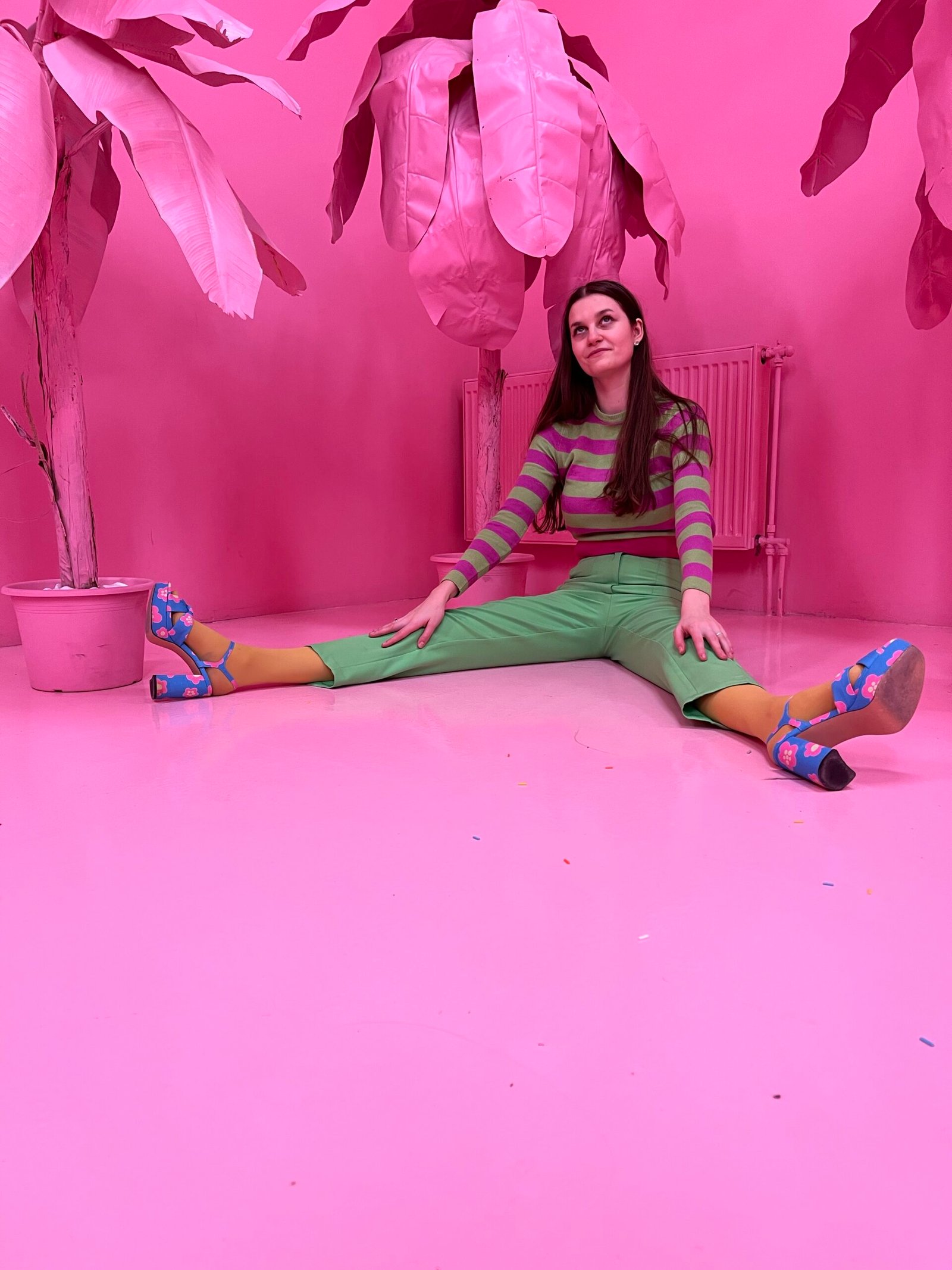 a girl wearing colorful clothes - green pants, pink and green blouse, blue sandals on heels, surrounded by pink palms