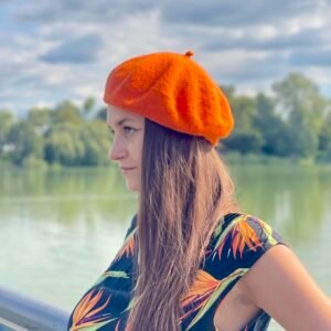 orange beret, side view, on a model with brown hair and black dress with orange flowers, a river and green trees in the background