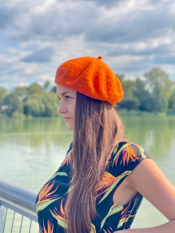 orange beret, side view, on a model with brown hair and black dress with orange flowers, a river and green trees in the background