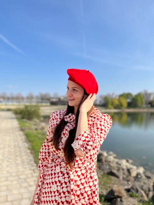 a woman wearing a red beret, red and white coat, she is smiling, an alley and a lake in the background