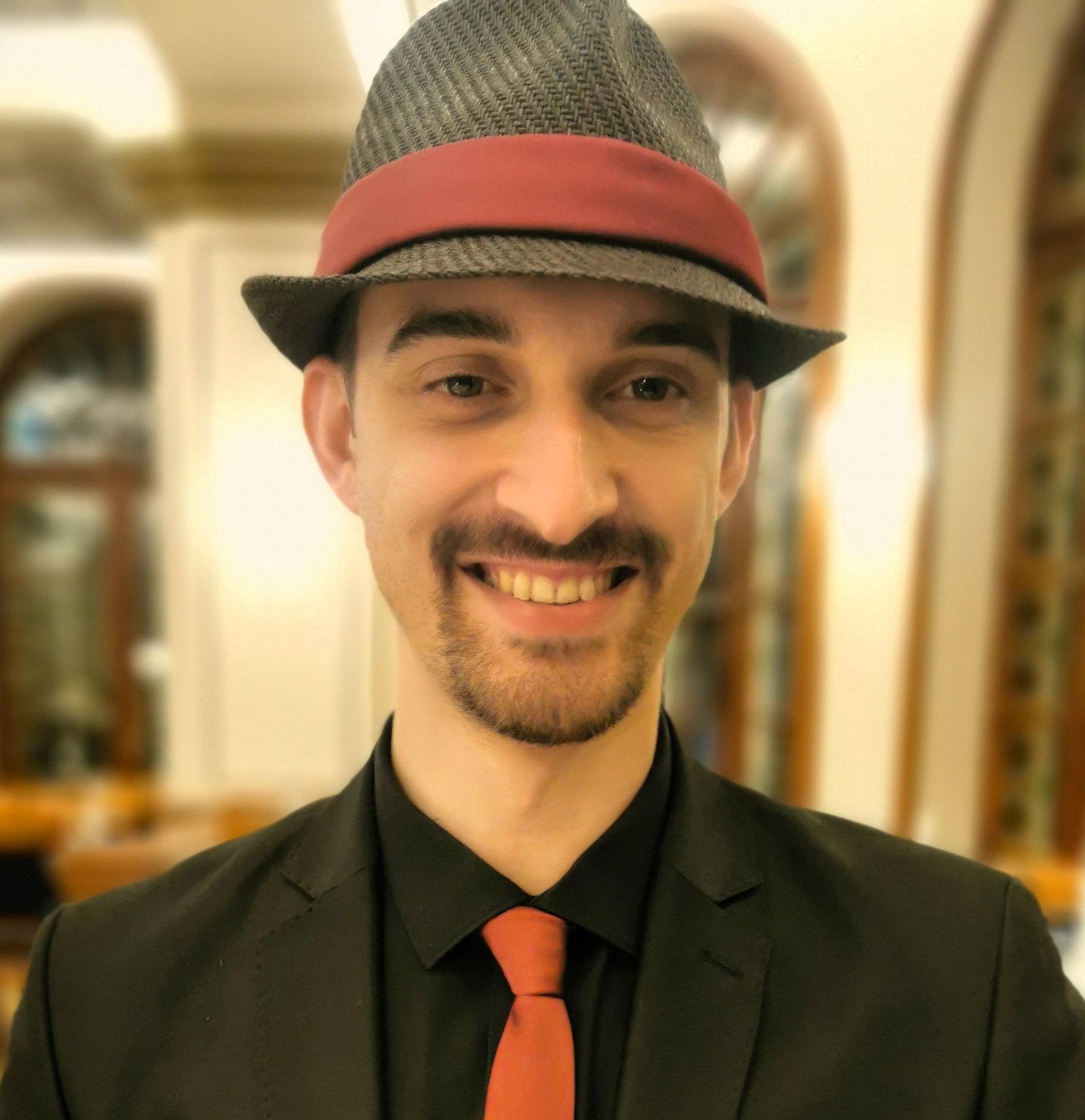 a man in black suit with a black hat and a dark red tie, smiling