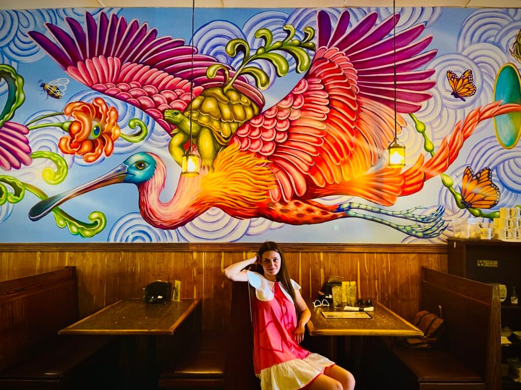 a girl sitting on a chair, a big wallpaper with the hottest colors of 2024 is behind here, presenting big colorful bird carrying a turtle