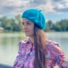 turquoise beret front view on a model with brown hair and colorful button shirt, a river and green trees in the background