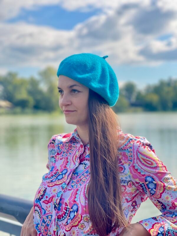 turquoise beret front view on a model with brown hair and colorful button shirt, a river and green trees in the background