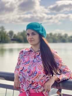 turquoise beret side view on a model with brown hair and colorful button shirt, a river and the clouds in the background