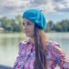 turquoise beret front view on a model with brown hair and colorful button shirt, a river and green trees in the background