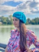 turquoise beret front view on a model with brown hair and colorful button shirt, a river and green trees in the background