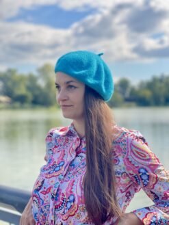 turquoise beret front view on a model with brown hair and colorful button shirt, a river and green trees in the background