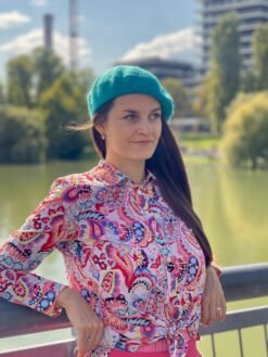 turquoise beret side view, a girl with brown hair wearing it, she is also wearing colorful, pinkish button shirt and looking at the horizon, a river and green trees in the background