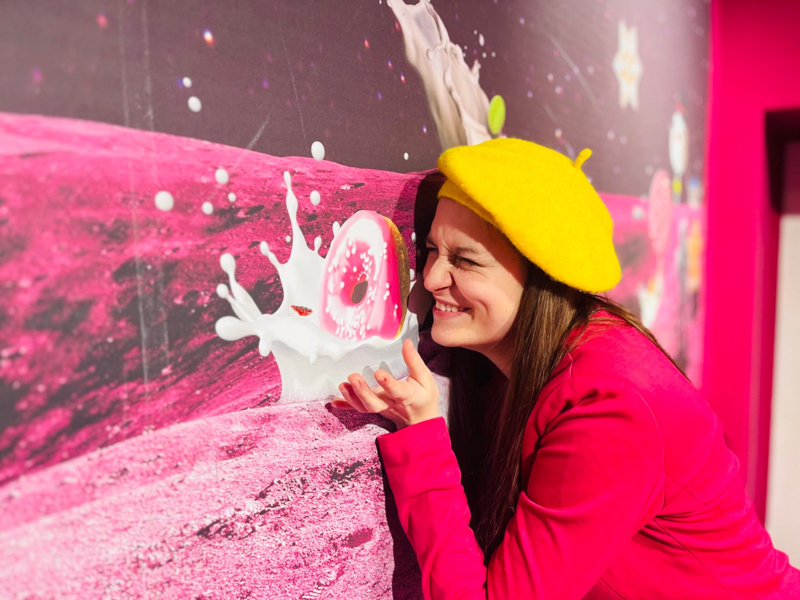 woman wearing a dopamine outfit featuring yellow beret and fuchsia blazer, she is smiling widely, the picture of a pink donut next to her