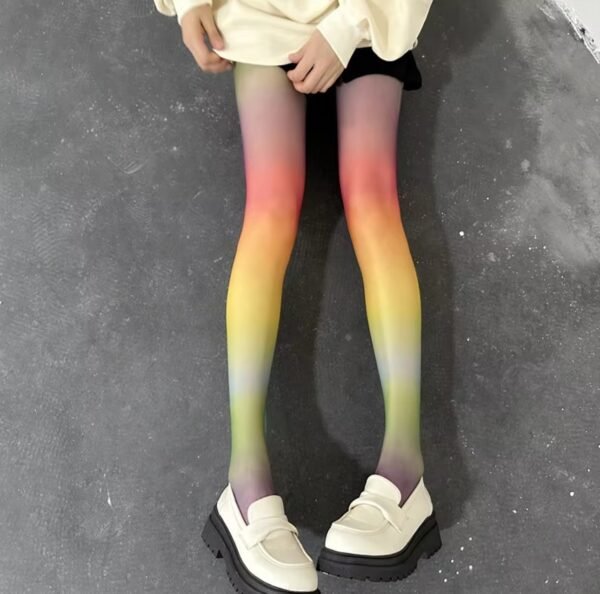 woman wearing rainbow tights, white loafers and a white jumper. she is sitting on the grey floor.