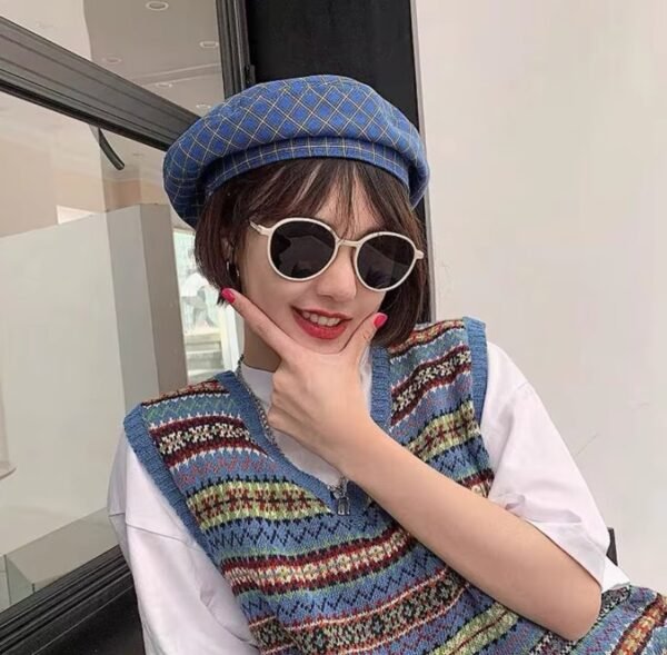 woman wearing blue plaid colorful beret and a crochet sweater and sunglasses, a window behind her