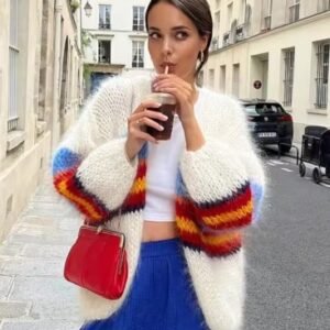 woman with brown hair wearing colorful fuzzy cardigan and blue pants, she is carrying a red purse and drinking a drink