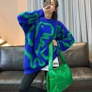 woman posing in colorful streetwear sweater and black pants. She is holding a green bag, a black sofa behind her.