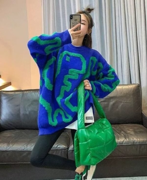 woman posing in colorful streetwear sweater and black pants. She is holding a green bag, a black sofa behind her.