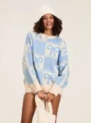 woman with brown curly hair wearing blue colorful sweater with ribbons and a white winter hat, she id holding a white bag and smiling