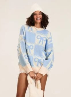 woman with brown curly hair wearing blue colorful sweater with ribbons and a white winter hat, she id holding a white bag and smiling