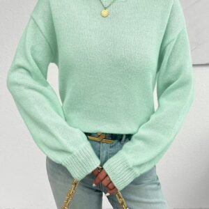 woman wearing mint sweater and bright jeans, she is holding a brown purse, white wall behind her