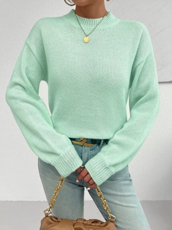 woman wearing mint sweater and bright jeans, she is holding a brown purse, white wall behind her