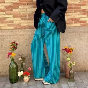 woman wearing green colorful baggy pants and black button shirt, wall and flowers next to her