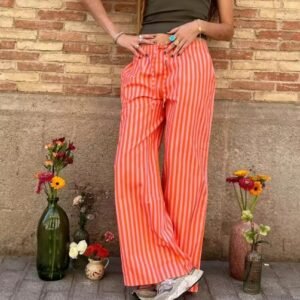 woman wearing orange colorful baggy pants and a grey top, grey wall and flowers next to her