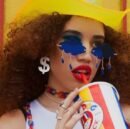 woman wearing colorful cloud shaped sunglasses and a yellow cowboy hat, she is drinking a drink through a red straw