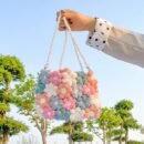 woman holding colorful floral handbag up in the air, blue sky and green tress in the background