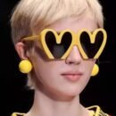 woman with short blonde hair wearing colorful heart shaped sunglasses and yellow earrings