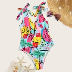 colorful V-neck one piece swimsuit lying on a beige carpet, flowers, suglasses and a sun hat next to it